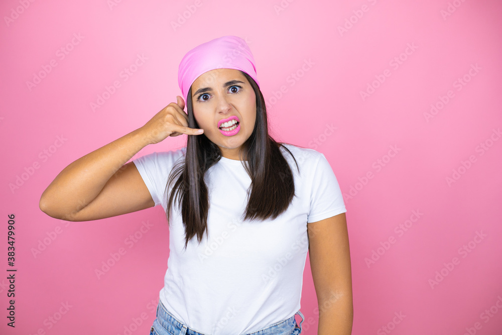 Young beautiful woman wearing pink headscarf over isolated pink background confused doing phone gesture with hand and fingers like talking on the telephone Young beautiful woman wearing pink headscarf over isolated pink background confused doing phone gesture with hand and fingers like talking on the telephone