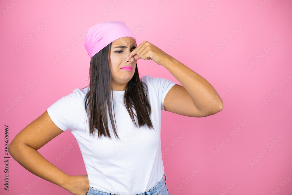 Young beautiful woman wearing pink headscarf over isolated pink background smelling something stinky and disgusting, intolerable smell, holding breath with fingers on nose