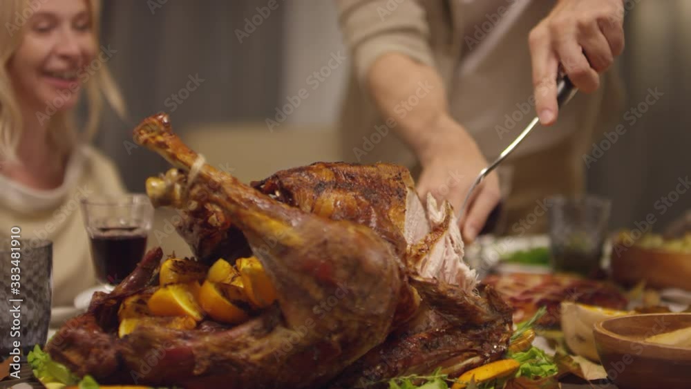 Unrecognizable mature man standing at dining table cutting roasted turkey for Thanksgiving day dinner