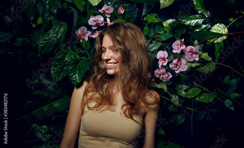 Beautiful young woman with smooth tanned skin laughs cheerfully while standing against the background of exotic plants and flowers. Natural cosmetics, skin care.