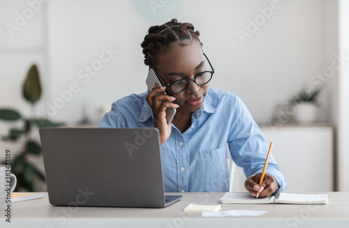 Black Female Office Worker Talking On Cellphone With Client And Taking Notes