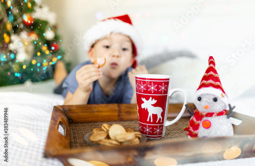 Joyful boy in bed eating cookies and drinking cacao. In Santa claus hat, New year tree on background. Christmas time 2021 photo