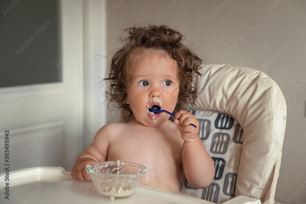 Adorable long haired toddler sitting in a baby high chair and eating porridge indoor. Adorable long haired toddler sitting in a baby high chair and eating porridge indoor.