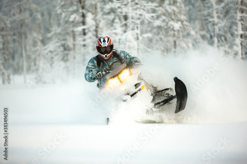 The rider in gear with a helmet makes a sharp turn on a snowmobile on a deep snow surface on a background of snowy landscaping nature and winter forest. photo