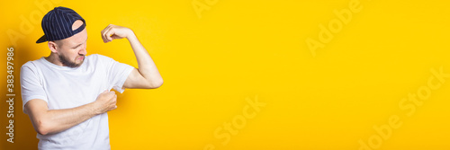 Young man in cap and t-shirt with sweaty and smelly armpits on yellow background