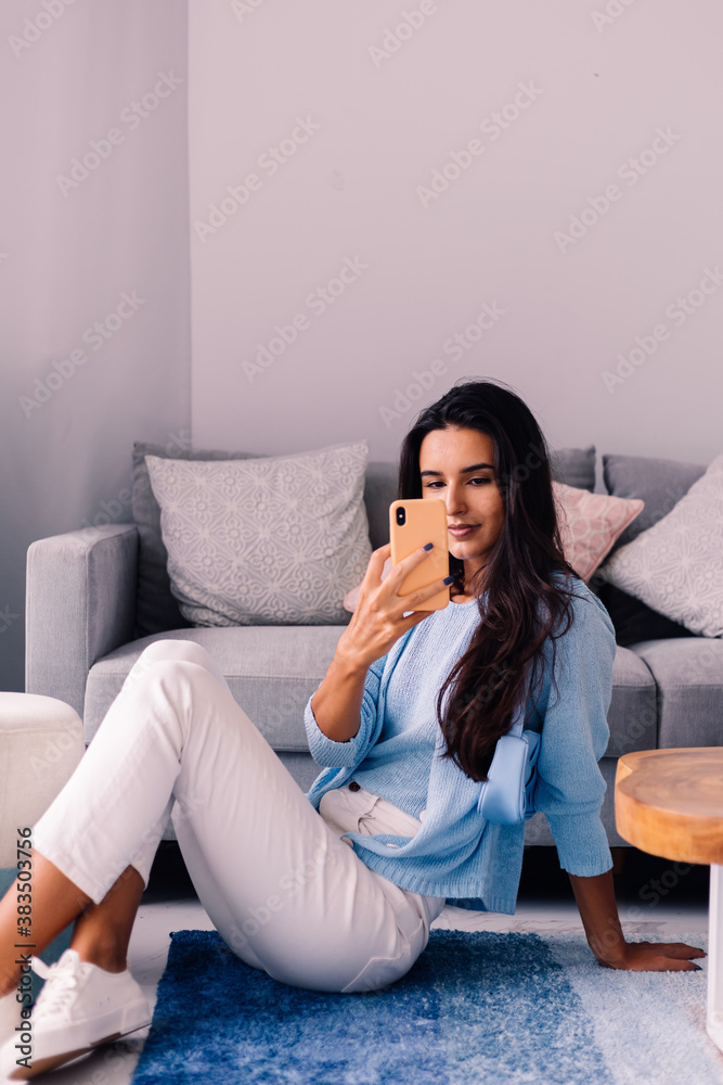 European fit brunette fashion blogger woman sits on floor in living room near sofa, wearing white jeans and blue sweather, and take photo selfie in mirror     