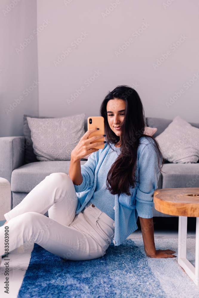 Obraz premium European fit brunette fashion blogger woman sits on floor in living room near sofa, wearing white jeans and blue sweather, and take photo selfie in mirror 