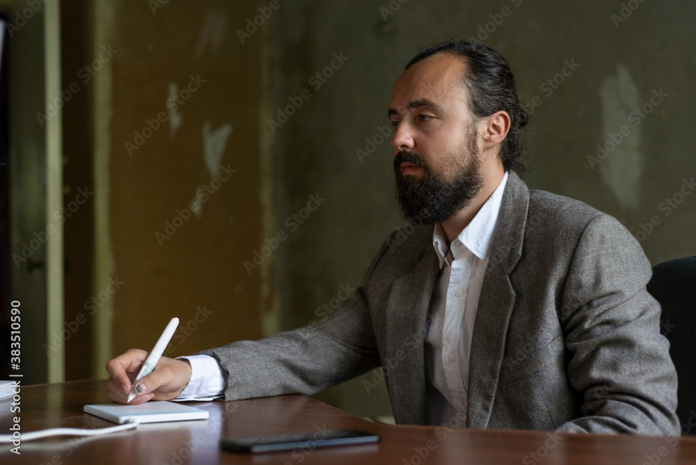 bearded young businessman in a business suit sitting at the computer table in the office. A computer mouse, writes a list in a graphical tablet. Office renovation bearded young businessman in a business suit sitting at the computer table in the office. A computer mouse, writes a list in a graphical tablet. Office renovation