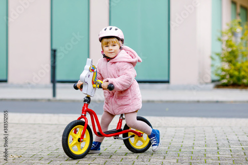Cute little toddler girl with helmet riding on run balance bike to daycare, playschool or kindergarden. Happy child having fun with learning on learner bicycle. Active kid on cold autumn day outdoors.