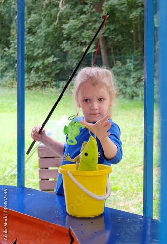 A little girl with blond long hair tied up in a ponytail is catching fish at a children's 