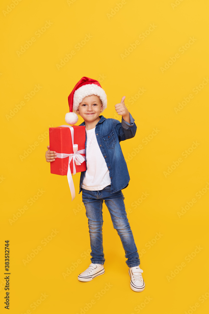 Portrait of a satisfied little child boy in christmas Santa hat. laughing isolated over yellow background. Holds a gift box. Preparing for the New Year holidays Portrait of a satisfied little child boy in christmas Santa hat. laughing isolated over yellow background. Holds a gift box. Preparing for the New Year holidays