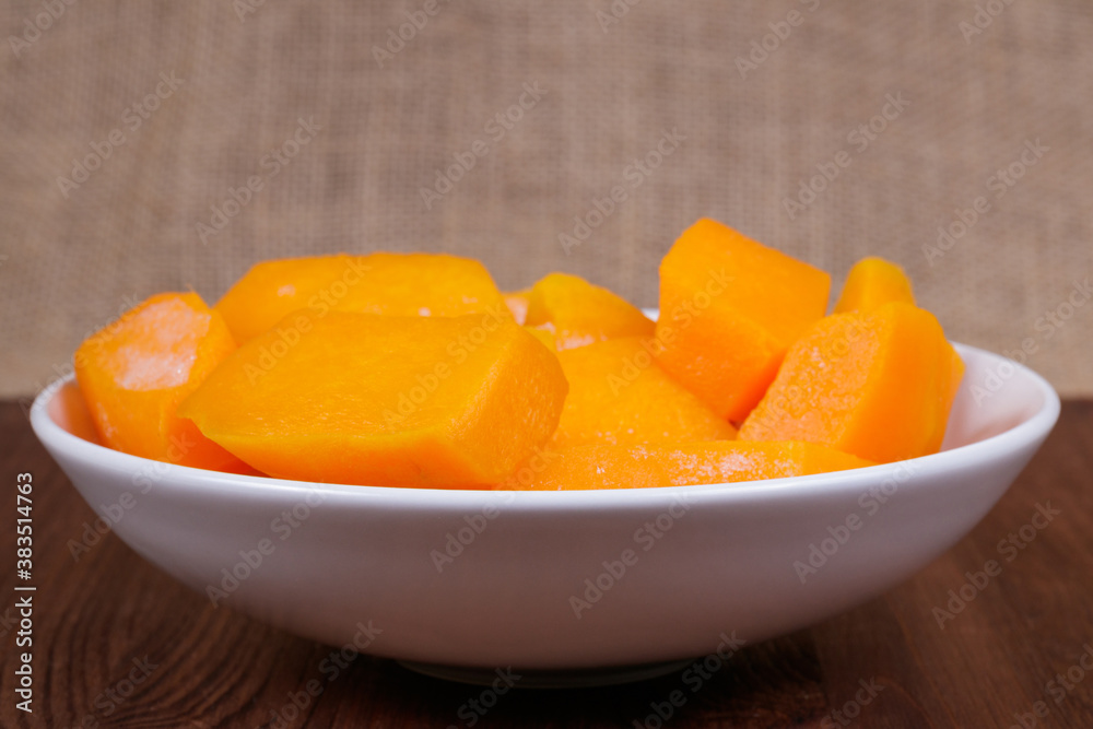 Pieces of pumpkin in the bowl at the wooden table and sackcloth background.