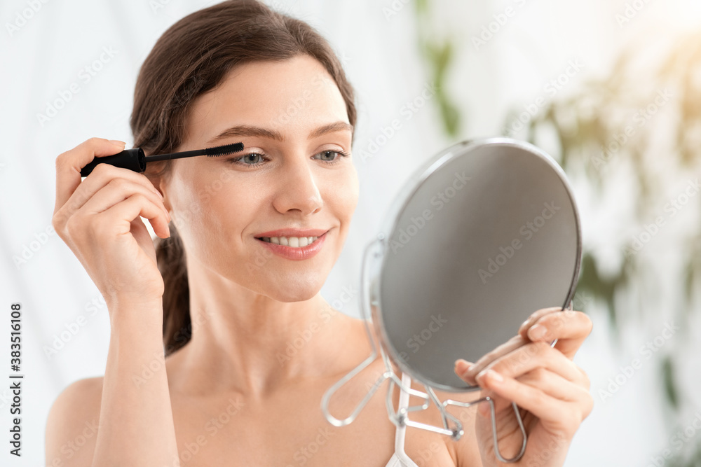 Smiling woman looking at mirror, applying mascara Smiling woman looking at mirror, applying mascara
