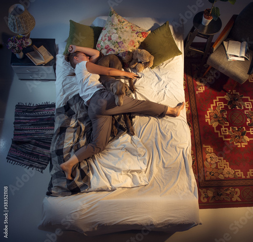 Portrait of a woman, female breeder sleeping in the bed with her dog at home. Top view. Dressed housekeeper sleeping after tiring work day. Holding his pet close to. Job, work, pets love concept.