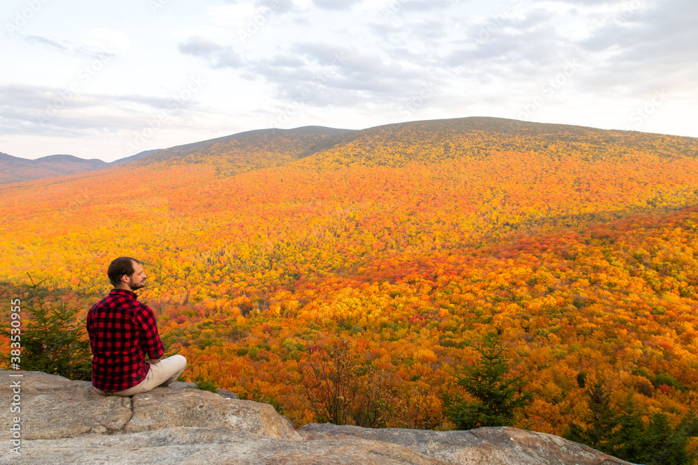 Young man with a lumberjack shirt sitting on the edge of a rock and admiring the beautiful autumnal colors in the Mont-Megantic national park, Canada Young man with a lumberjack shirt sitting on the edge of a rock and admiring the beautiful autumnal colors in the Mont-Megantic national park, Canada