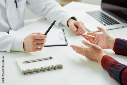 A close photo of the hands of a female patient and a doctor at an appointment in a hospital. A notebook with a pen, a clipboard, a laptop are lying on a white desk in a doctor's office.