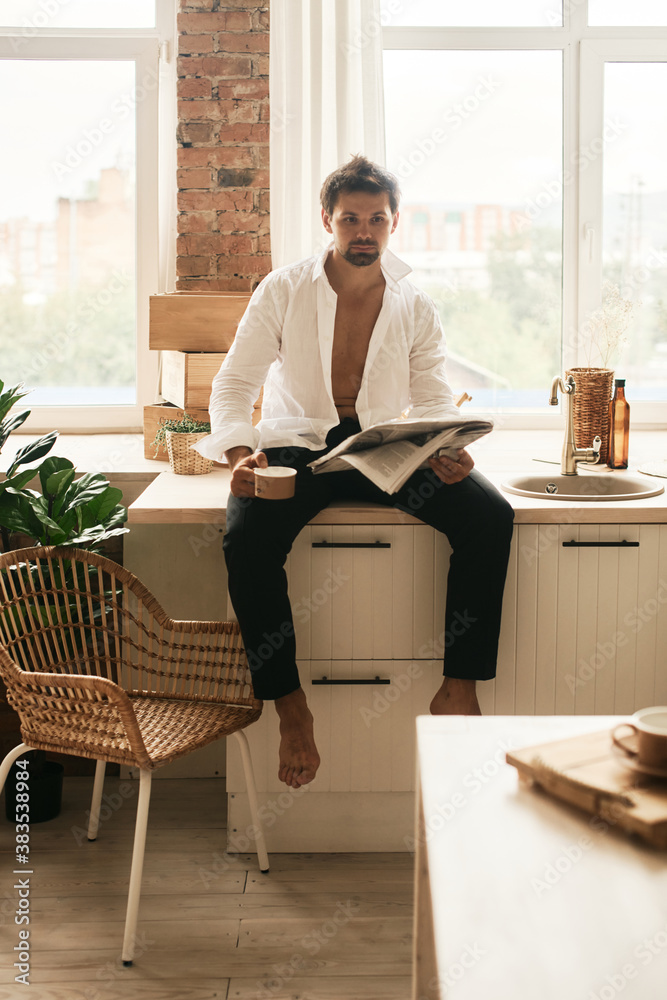 European man in his flat. Sitting in the living room. Bronze skin. Dark eyes and hair. Beard. Light colored interior. Casual clothes style. Weekend at home. Domestic atmosphere. Cosiness and comfort