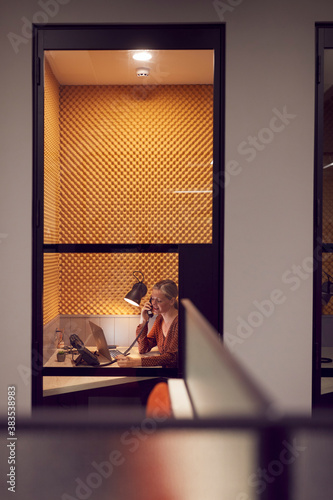 Businesswoman Working Late In Individual Office Cubicle On The Phone Using Laptop