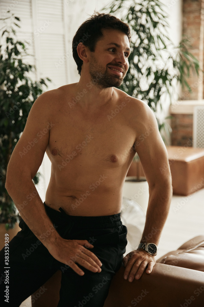 Obraz premium European man in his flat. Sitting in the living room. Bronze skin. Dark eyes and hair. Beard. Light colored interior. Casual clothes style. Weekend at home. Domestic atmosphere. Cosiness and comfort
