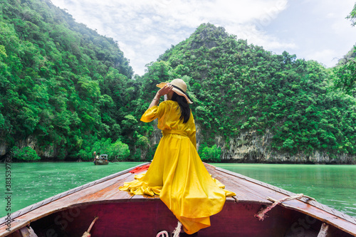 Woman wearing a long yellow dress, sitting on local long-tail boat, holding her hands on hat, she travels to Hong Island, beautiful sea in Krabi, Thailand.