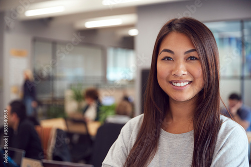 Head And Shoulders Portrait Of Smiling Young Asian Businesswoman Working In Busy Modern Office