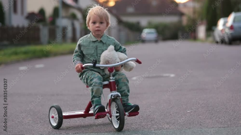 Toddler child, blond boy, riding tricycle in a village small road on sunset with teddy bear Toddler child, blond boy, riding tricycle in a village small road on sunset with teddy bear