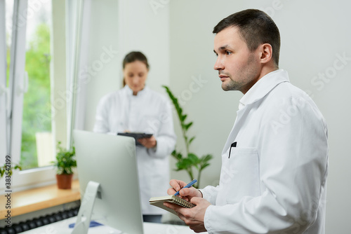 A caucasian doctor is doing notes with a pen near a female therapist with a clipboard near an all in one computer in a hospital.