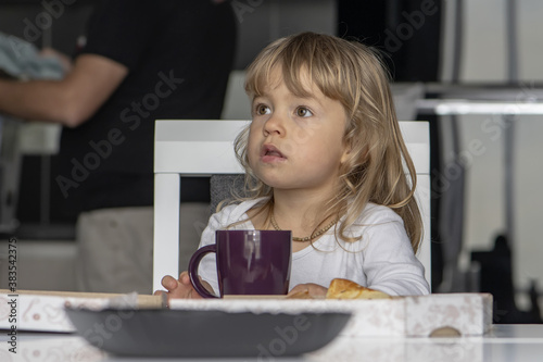 A little girl is sitting at the kitchen table with a mug and a pizza box in front of her.
