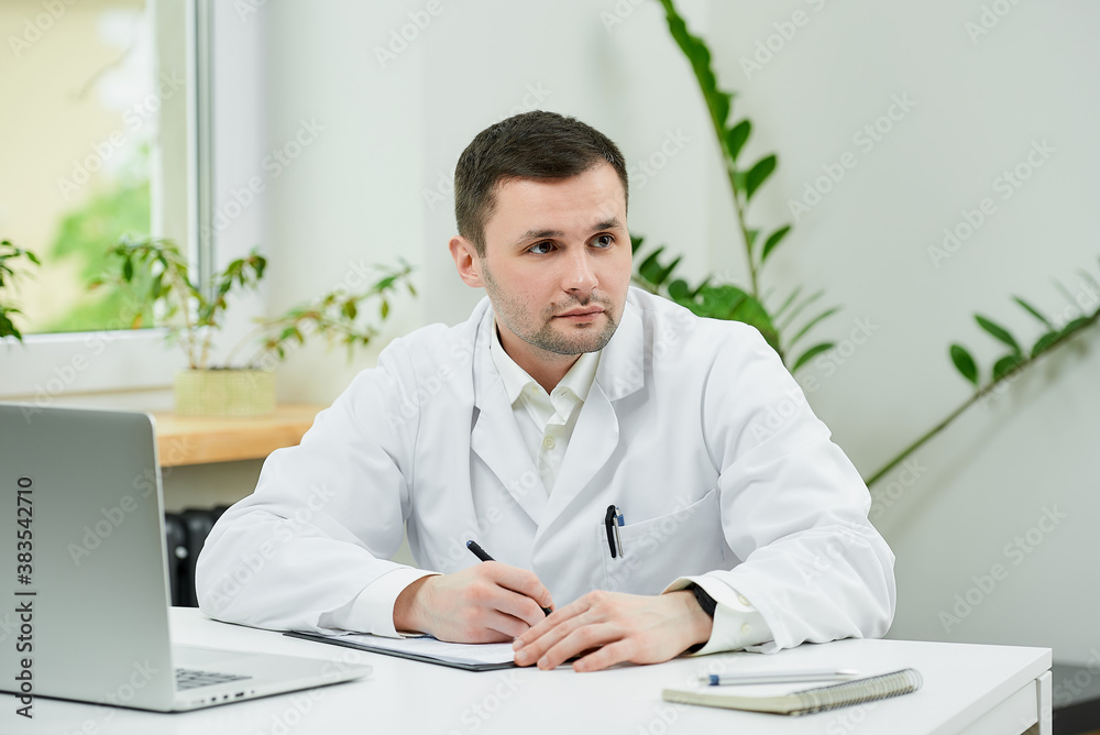 Fototapeta premium A caucasian doctor in a white lab coat is filling out a patient card with a diagnosis and thinking in a hospital. A therapist near a laptop is waiting for a patient in a doctor's office.