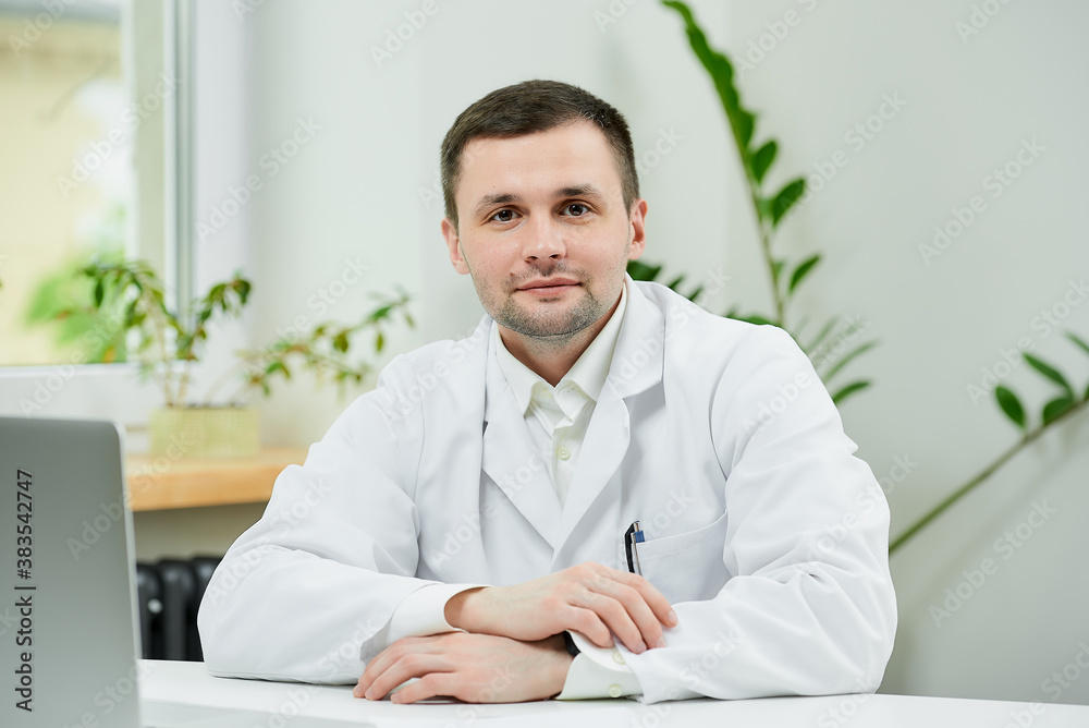 A kind caucasian doctor in a white lab coat is sitting arms crossed in a hospital. A therapist near a laptop is waiting for a patient in a doctor's office.
