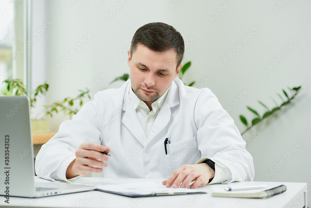 A serious caucasian doctor in a white lab coat is sitting at his desk and examining the patient’s medical history in a hospital. A therapist near a laptop in a doctor's office. A serious caucasian doctor in a white lab coat is sitting at his desk and examining the patient’s medical history in a hospital. A therapist near a laptop in a doctor's office.