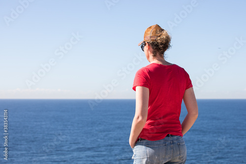 A woman in a red top t shirt denim shorts from behind happy success freedom against blue sky on bright sunny summer day outdoors