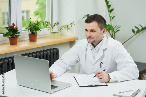 A happy caucasian doctor in a white lab coat is listening to a patient on an online meeting on a computer in a hospital. A therapist is sitting at a desk with a laptop in a doctor's office.