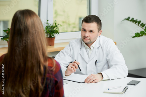 Obraz na płótnie A serious caucasian doctor in a white lab coat is sitting at a desk near a female patient in a doctor's office. A woman with long hair at a doctor's appointment in a hospital. w salonie