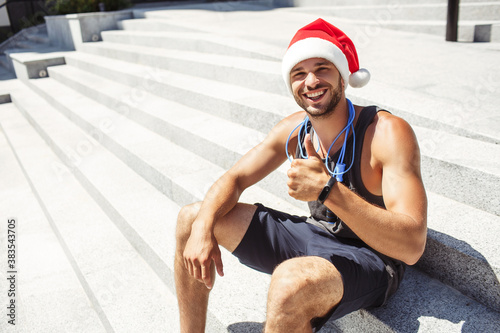 Young man exercising outside. Guy wearing christmas santa hat and show big thumb up. Like symbol. Strong powerful athletist sit alone outisde on steps under the sun. photo