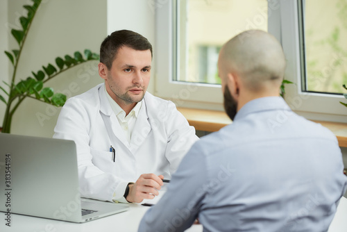 A caucasian doctor in a white lab coat is sitting at a desk and carefully listening to a story of a bald male patient in a hospital. A man with a beard at an appointment in a doctor s office.