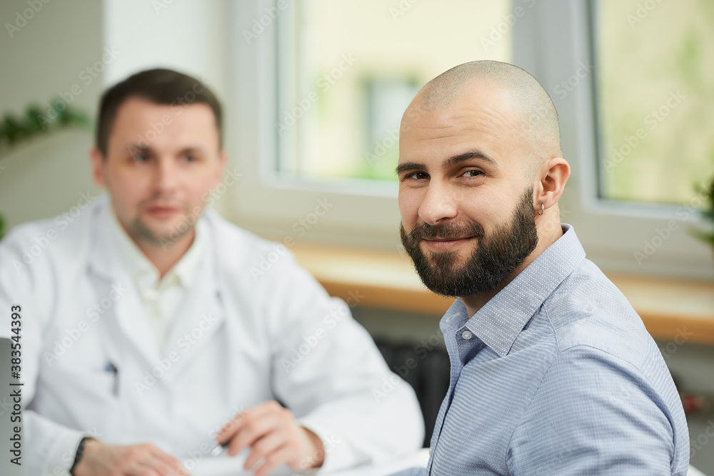 A bald male patient is smiling near a caucasian doctor in a white lab coat in a hospital. A man with a beard at an appointment in a doctor's office.