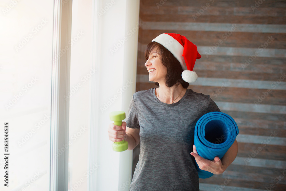 Adult fit slim woman has workout at home. New year or Christmas time period.  Well-built adult senior woman resting after workout in room. Hold green  dumbbell and blue yoga mat. Stock Photo