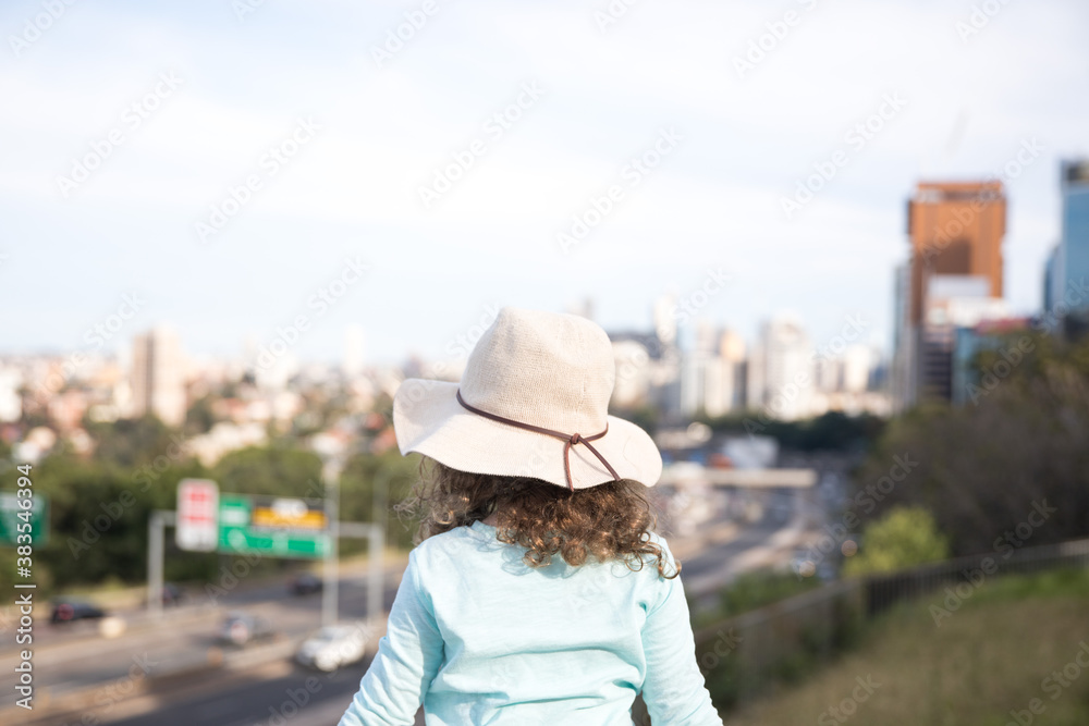 View of a young girl child from behind wearing a broad rim sun hat blue top with her arms outstretched in excitement looking out over the city landscape cityscape Sydney NSW