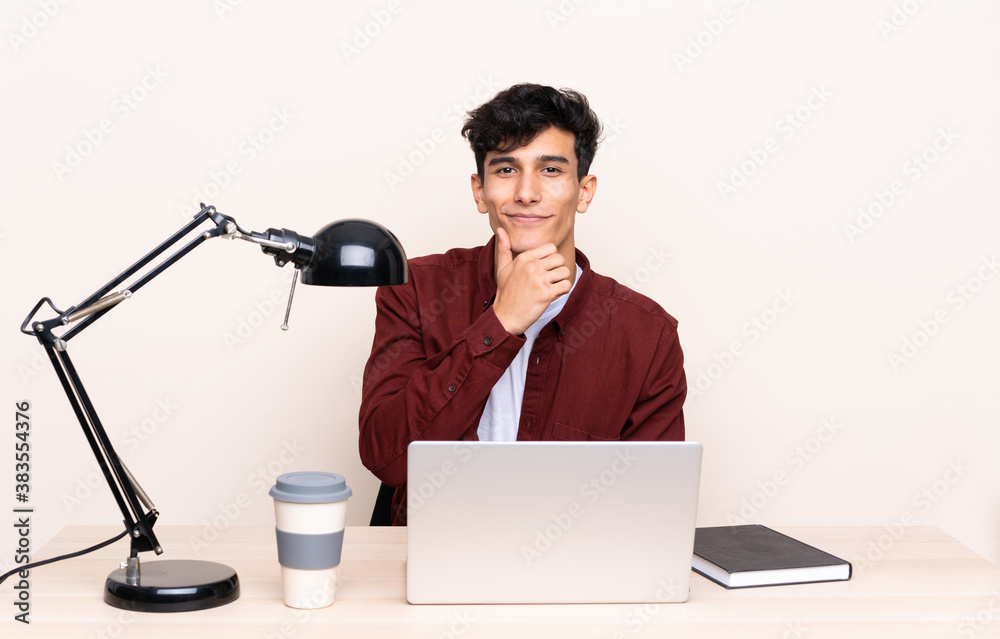 Young Argentinian man in a table with a laptop in his workplace laughing