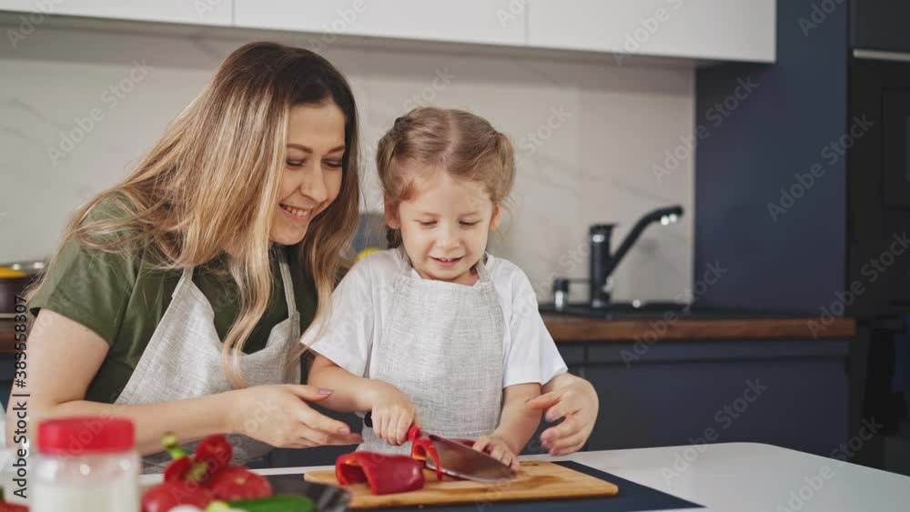 happy young mother looks at little daughter trying to cut ripe red bell pepper for salad on wooden board at table in light modern kitchen happy young mother looks at little daughter trying to cut ripe red bell pepper for salad on wooden board at table in light modern kitchen