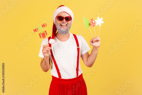 Happy positive gray bearded man in santa claus costume holding paper cards on sticks, celebrating christmas and congratulating with new year. Indoor studio shot isolated on yellow background