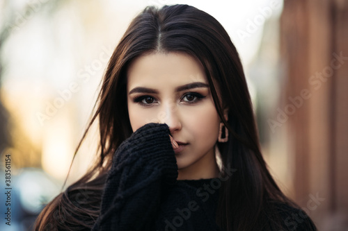 Close up lifestyle portrait of young indian woman is cold standing on the street cloudy weather, dressed in black pullover and looking at the camera. 