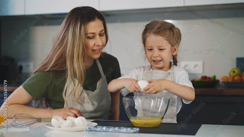 cheerful woman looks at pretty little girl in apron cracking chicken egg into glass bowl at table in light modern kitchen at home cheerful woman looks at pretty little girl in apron cracking chicken egg into glass bowl at table in light modern kitchen at home