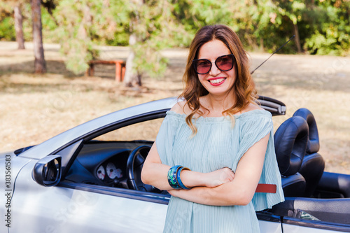 Attractive woman leaning and posing at convertible car. Happy pretty girl. Outdoor fashion portrait. Wearing elegant dress, straw hat and sunglasses. Looking at camera. 