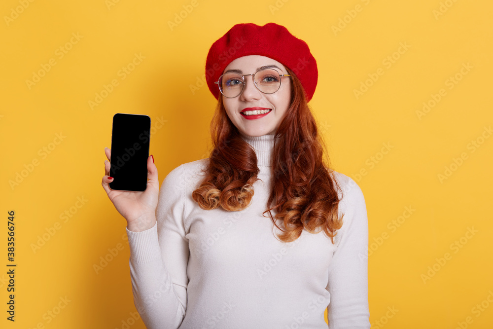 Image of smiling red haired girl wearing red beret and white shirt, girl with red lips holding smart phone with blank screen in hands, isolated on yellow. Copy space or promotion text. Image of smiling red haired girl wearing red beret and white shirt, girl with red lips holding smart phone with blank screen in hands, isolated on yellow. Copy space or promotion text.