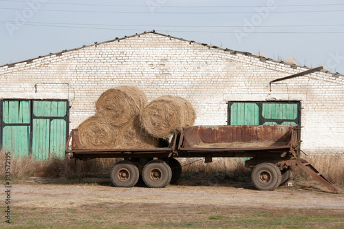 Round skeins of hay on an old rusty trailer to a tractor against the background of a white brick building with a green gate