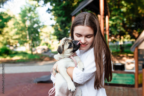 Happy dog ​​and girl. The girl is playing with a dog. Young woman walking with a pug dog in summer park. Portrait of a pug. Portrait of a beautiful pug puppy. The dog is lying on the ground. © Alex