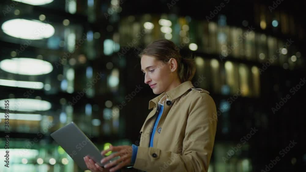Side view slow motion shot of young businesswoman in trench coat opening laptop, using pc and looking away thoughtfully standing in dark city street at windy night Side view slow motion shot of young businesswoman in trench coat opening laptop, using pc and looking away thoughtfully standing in dark city street at windy night