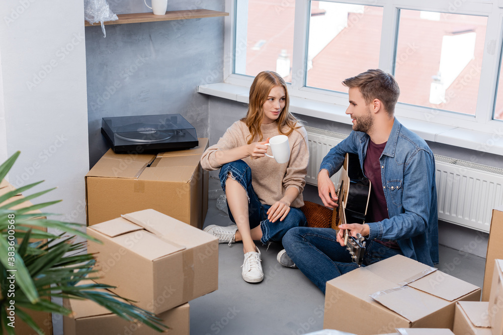 selective focus of young man sitting on floor playing acoustic guitar near woman with cup and carton boxes selective focus of young man sitting on floor playing acoustic guitar near woman with cup and carton boxes